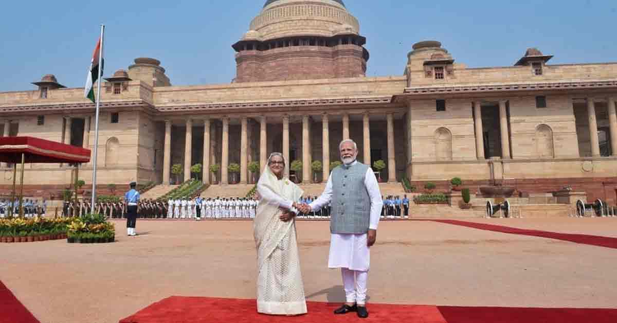 Indian Prime Minister Narendra Modi and Bangladesh Prime Minister Sheikh Hasina are seen smiling and shaking hands at a formal event. Both leaders are dressed in traditional attire; Modi is in a white kurta with a beige jacket, while Hasina is in a colorful saree."